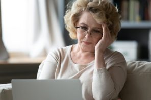 Mulher idosa de blusa bege e óculos de grau sentada no sofá com notebook no colo, apoiando uma das mãos na cabeça e demonstrando frustração ao se deparar com o erro 404 em uma página da web.