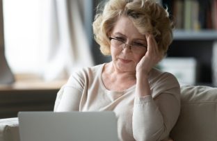 Mulher idosa de blusa bege e óculos de grau sentada no sofá com notebook no colo, apoiando uma das mãos na cabeça e demonstrando frustração ao se deparar com o erro 404 em uma página da web.