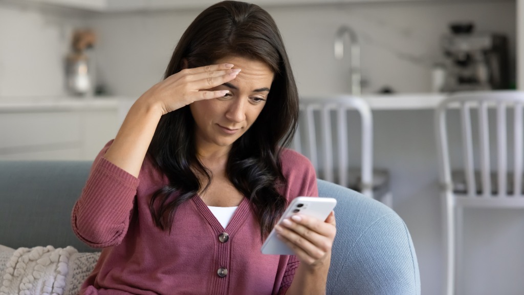 Mulher jovem com blusa vinho sentada no sofá da sala de estar, com uma das mãos na testa e expressão de espanto devido a problemas no sinal do celular.
