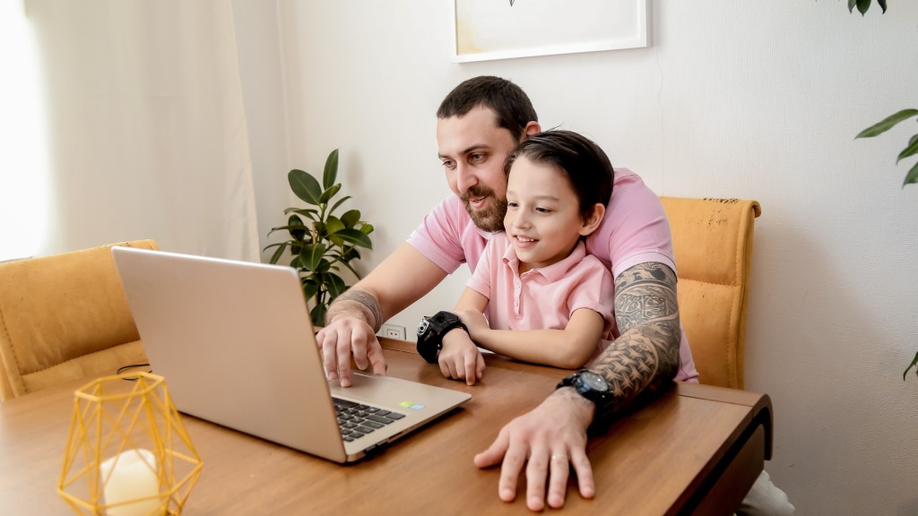Pai e filho, ambos com camisas rosa clara, sentados à mesa olhando a tela do notebook, simbolizando o bloqueio de sites para uma navegação segura.