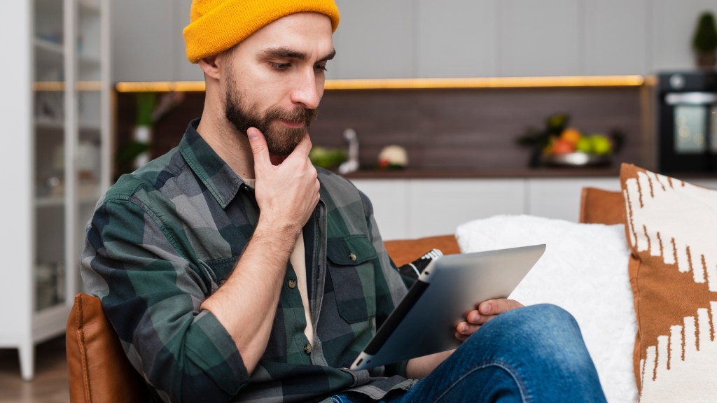 Homem com camisa xadrez preta, verde e cinza, calças jeans e touca amarela, sentado no sofá com tablet na mão, apoiando a outra mão no queixo, com expressão de dúvida sobre como escolher o melhor provedor de internet.