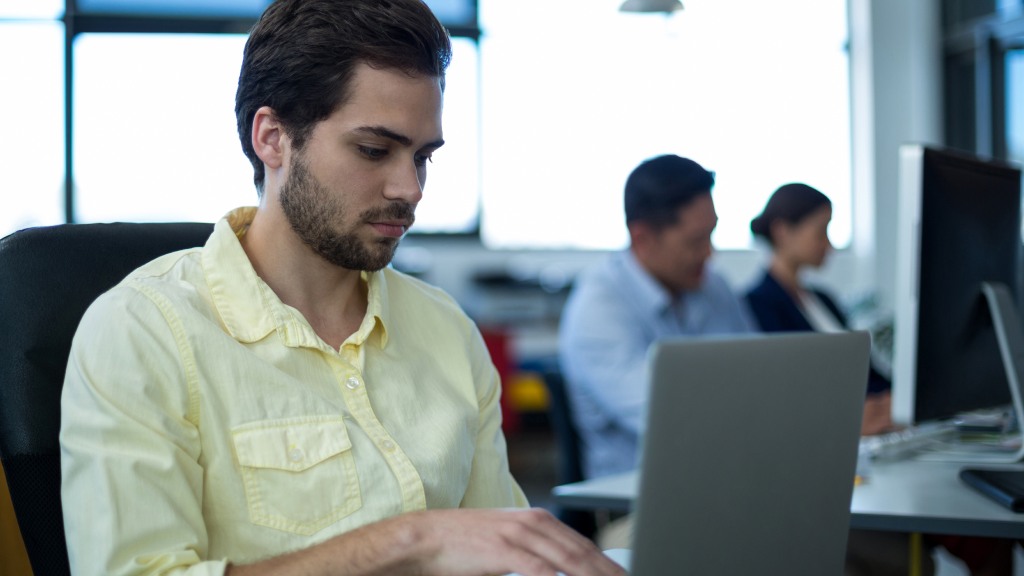 Homem jovem com camisa de botões amarela, sentado à mesa de computador no escritório, trabalhando no notebook conectado à internet fibra empresarial, com dois colegas desfocados ao fundo.