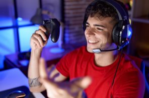 Rapaz gamer com camiseta vermelha, headphones pretos e controle de videogame na mão direita, sorrindo durante a partida após monitorar quantos Mega tem sua internet.