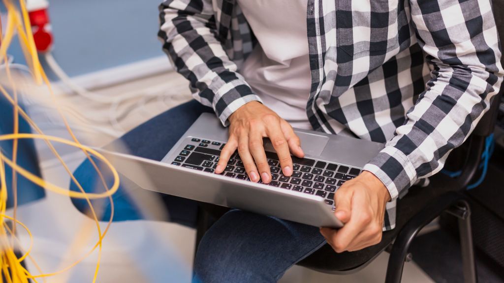 Homem mostrado do peito para baixo usando camisa xadrez preta, branca e cinza e calças jeans, notebook no colo e cabos amarelos, representando provedor de internet.