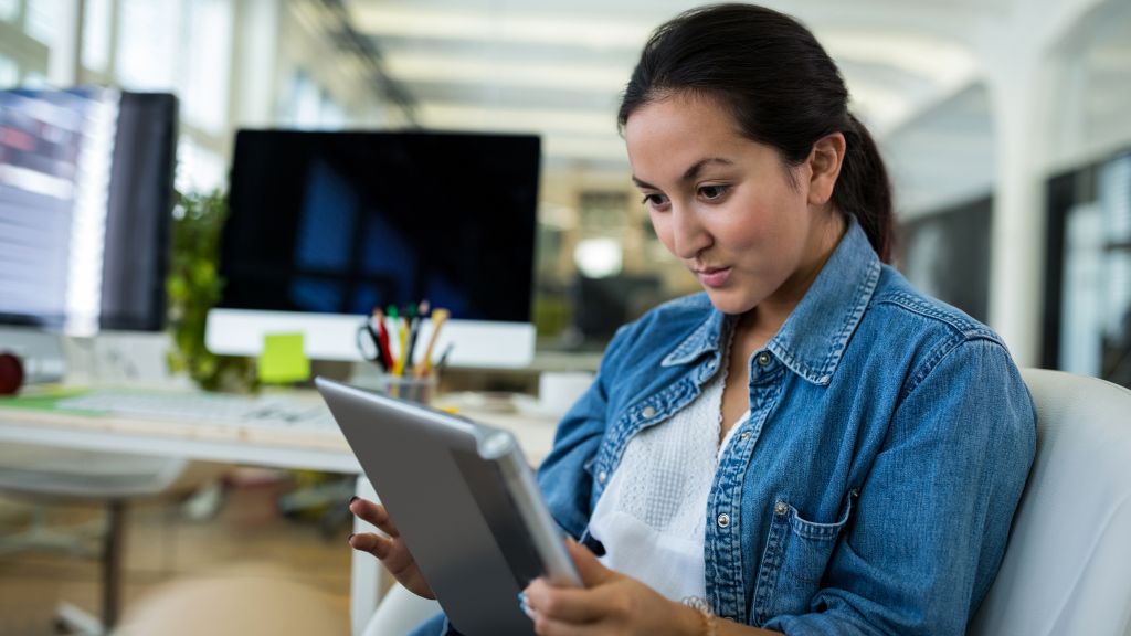 Mulher jovem com camisa jeans, concentrada, sentada em poltrona do escritório, usando tablet conectado à internet fibra corporativa.
