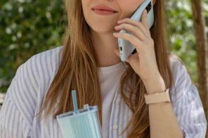 Mulher jovem com blusa listrada lilás e branca, segurando um copo azul grande e com o celular no ouvido, fazendo ligação para outro DDD.