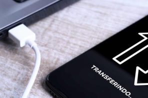 Celular conectado via USB a um notebook sobre uma mesa, representando a dúvida sobre como transferir arquivos do celular para o PC.