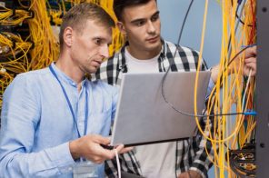 Dois homens de pé manuseando fios amarelos de internet, um com camisa azul clara segurando notebook e outro com camisa xadrez amarela, representando profissionais de uma empresa provedora de internet.