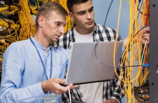 Dois homens de pé manuseando fios amarelos de internet, um com camisa azul clara segurando notebook e outro com camisa xadrez amarela, representando profissionais de uma empresa provedora de internet.