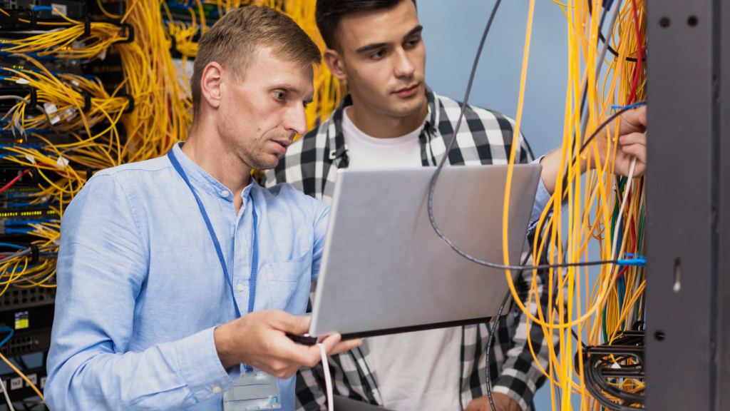 Dois homens de pé manuseando fios amarelos de internet, um com camisa azul clara segurando notebook e outro com camisa xadrez amarela, representando profissionais de uma empresa provedora de internet.