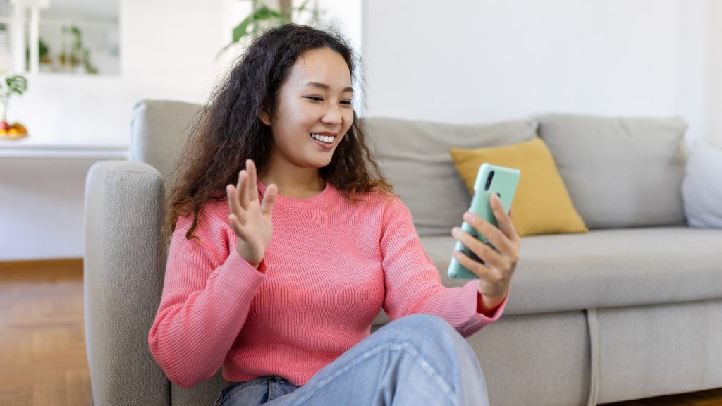 Moça com blusa rosa e calças jeans, sorrindo, sentada no chão da sala em frente a um sofá cinza, segurando o celular e fazendo um gesto de cumprimento, aproveitando os benefícios da portabilidade de chip com a Desktop.