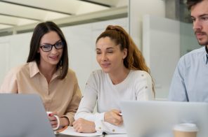 Três colegas sentados à mesa de trabalho, concentrados, olhando para a tela do notebook conectado à internet fibra empresarial: um rapaz com camisa social azul clara, uma mulher com blusa branca e outra com camisa social marrom clara.
