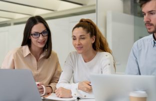 Três colegas sentados à mesa de trabalho, concentrados, olhando para a tela do notebook conectado à internet fibra empresarial: um rapaz com camisa social azul clara, uma mulher com blusa branca e outra com camisa social marrom clara.