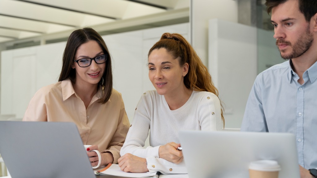 Três colegas sentados à mesa de trabalho, concentrados, olhando para a tela do notebook conectado à internet fibra empresarial: um rapaz com camisa social azul clara, uma mulher com blusa branca e outra com camisa social marrom clara.