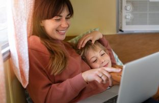 Mãe e filho deitados juntos: ela com blusa vermelha, ele com blusa laranja deitado sobre seu braço, ambos sorrindo, enquanto ela aponta para a tela do notebook, simbolizando acesso seguro a sites.