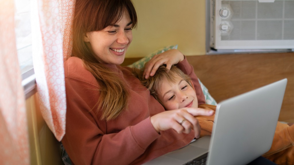 Mãe e filho deitados juntos: ela com blusa vermelha, ele com blusa laranja deitado sobre seu braço, ambos sorrindo, enquanto ela aponta para a tela do notebook, simbolizando acesso seguro a sites.