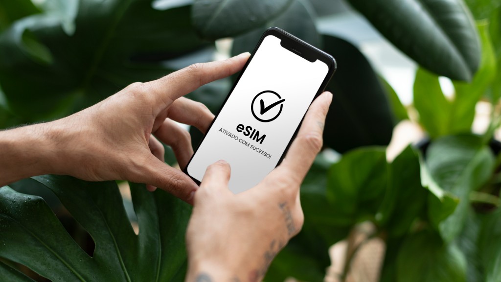Mãos segurando um celular que exibe na tela o texto “eSIM ativado com sucesso” e um ícone de check no topo, plantas verdes ao fundo, simbolizando a portabilidade de chip concluída com sucesso.
