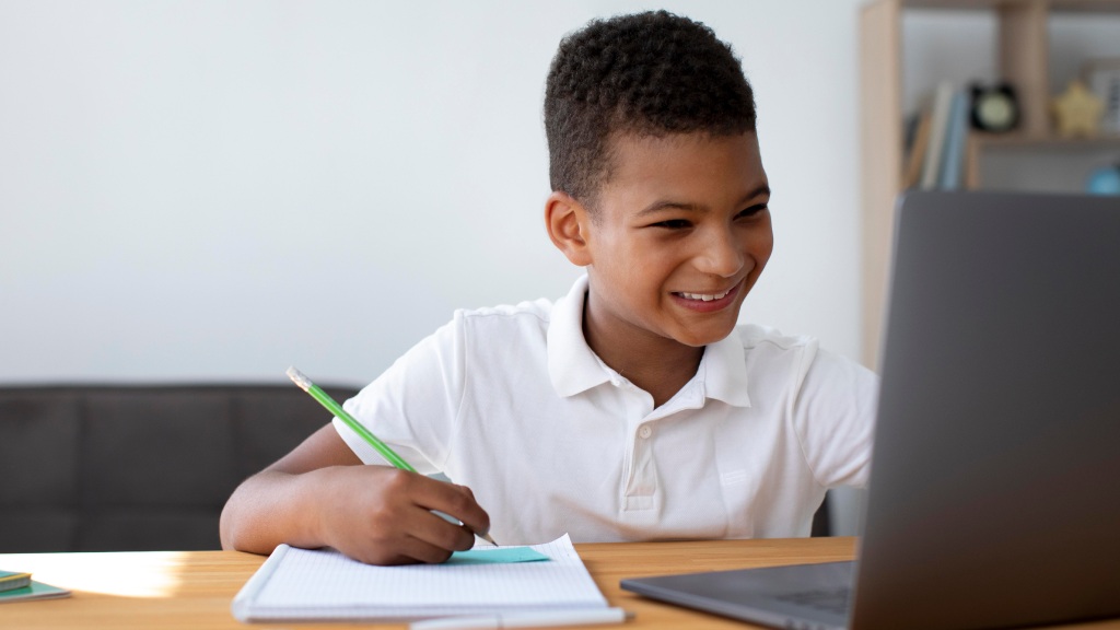 Menino com camisa branca, sentado à escrivaninha com notebook, caderno e lápis na mão, sorrindo enquanto navega com segurança graças ao bloqueio de sites.