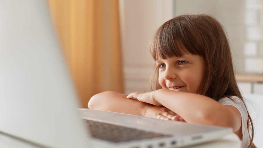 Menina pequena com franja, sorrindo, sentada à mesa do notebook e olhando para a tela, simbolizando navegação segura com bloqueio de sites.