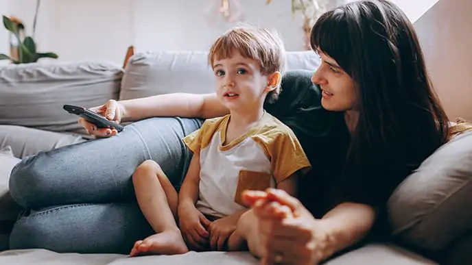 Conheça os principais canais infantis e os pacotes da Desktop em que eles estão inseridos. Uma mãe e o seu filho pequeno, deitados no sofá. Ela segura o controle remote da televisão e a criança assiste a um dos canais infantis da TV Desktop.