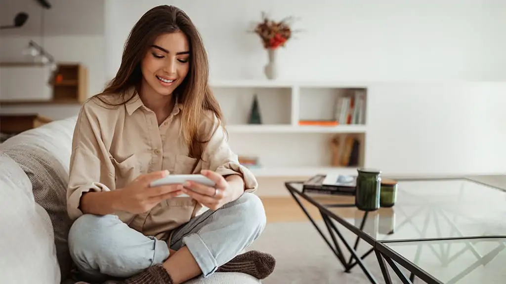 Usar o seu avatar durante uma conversa no WhatsApp pode ser muito divertido. Uma mulher, sentada no sofá, segurando o celular e enviando uma avatar.