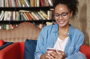 Uma adolescente, sentada no sofá lendo no aplicativo do Skeelo. Na imagem, ao fundo, tem um estante com diversos livros