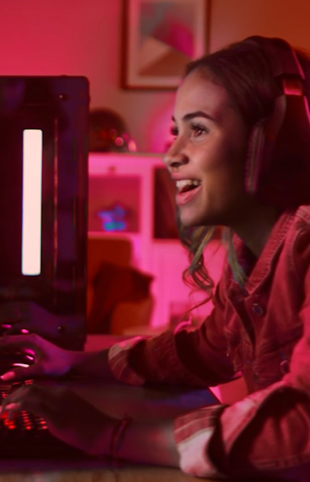 Uma moça, jogando online no computador. Ela usa um fone de ouvido e está concentrada na partida